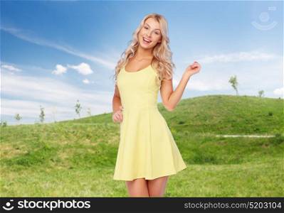 fashion, summer and people concept - happy smiling beautiful young woman posing in yellow dress over blue sky and green field background. smiling beautiful young woman in summer dress