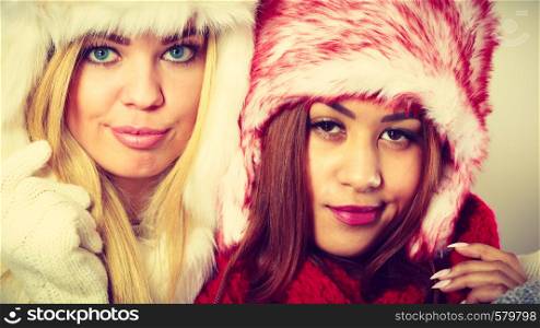 Fashion winter outfit concept. Two girls blonde and mulatto in warm red white clothing portrait. Attractive women wearing fur caps, scarfs, gloves.. Two girls in warm winter clothing portrait.