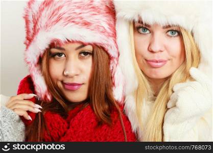 Fashion winter outfit concept. Two girls blonde and mulatto in warm red white clothing portrait. Attractive women wearing fur caps, scarfs, gloves.. Two girls in warm winter clothing portrait.