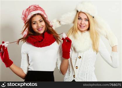 Fashion winter outfit concept. Two happy smiling girls blonde and mulatto in warm red white clothing portrait. Attractive women wearing fur caps, scarfs, gloves.. Two smiling girls in warm winter clothing.