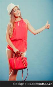 Fashion woman in hat and red shirt with handbag.. Cute attractive young woman girl in red shirt and straw hat with handbag in studio on blue showing thumb up gesture. Summer female fashion vogue.
