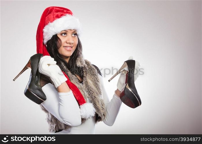 Fashion woman in santa hat with high heels shoes.. Fashion mixed race african american woman in santa claus hat holding black high heels shoes. Gorgeous young girl in studio on gray. Female fashion.