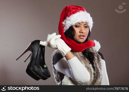 Fashion woman in santa hat with high heels shoes.. Fashion mixed race african american woman in santa claus hat holding black high heels shoes. Gorgeous young girl in studio on gray. Female fashion.