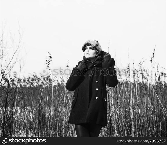 Fashion woman wearing a winter coat and fur cap and she posing front of the reeds, cold rainy weather, waist up, black and white image