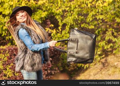 Fashion woman with bag outside. Beauty fashionable young woman having fun in park. Model wearing modern clothes in motion playing with her black bag outdoor.
