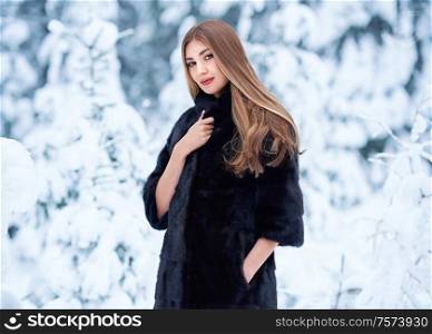 Fashion young woman in the winter forest. Young elegant model in black fur coat in nature. Blonde girl with long smooth hair