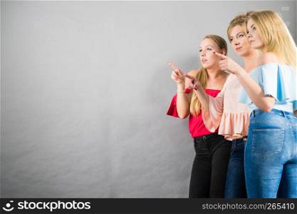 Fashionable adult female friends models pointing at grey copy space. Studio shot. Three fashionable females pointing