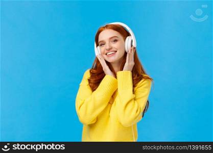 Fashionable attractive, carefree redhead woman in yellow sweater listening music with headphones, smiling tilting head joyfully, receive new earphones as christmas gift, standing blue background.. Fashionable attractive, carefree redhead woman in yellow sweater listening music with headphones, smiling tilting head joyfully, receive new earphones as christmas gift, standing blue background