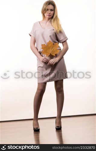 Fashionable autumn long hair blonde girl wearing beige dress high heels holding autumnal dry brown maple leaf in hand. Autumn, season and fashion concept.. Woman holding maple leaf in hand