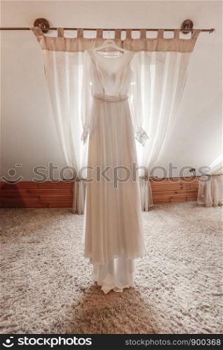 Fashionable beautiful classic lace silk wedding dress hanging on hanger in hotel wooden room. morning preparation wedding concept. vintage wedding gown.. stylish and beautiful wedding dress. classic lace silk wedding dress hanging on hanger in hotel wooden room. morning preparation wedding concept.