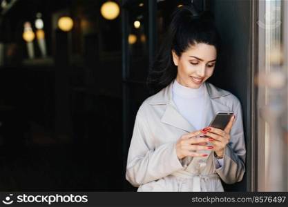 Fashionable beautiful woman with dark hair tied in pony tail dressed in white elegant coat holding cell phone looking into screen with delightful expression. Young pretty model communicating online