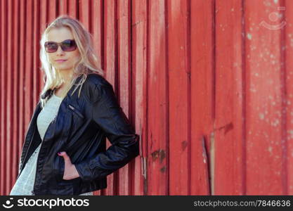 Fashionable beautiful young blond wearing a leather jacket and leans against the red wall