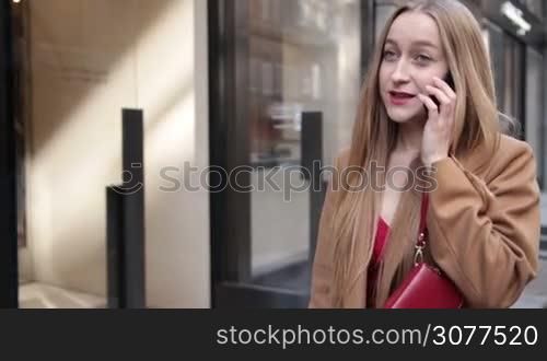 Fashionable blond woman with amazing long hair and deep blue eyes talking on smart phone while walking in the street. Stunning young female with red clutch wallet and camel coat talking on cell phone while going to business meeting. Slow motion.