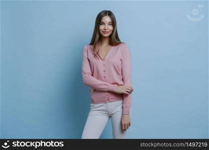Fashionable brunette Caucasian woman dressed in pink jumper and white trousers, going to work, applies makeup, smiles pleasantly, isolated on blue background. People, style, lifestyle concept