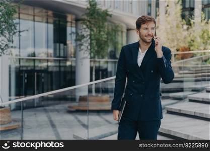 Fashionable businessman uses modern technologies has telephone conversation holds electronic gadget poses outdoor near office center. Successful economist makes phone call focused into distance. Fashionable unshaven businessman uses modern technologies has telephone conversation holds gadget