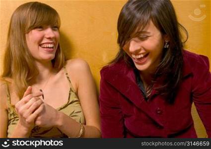 Fashionable Caucasian Girlfriends Sitting Against A Yellow Wall And Laughing