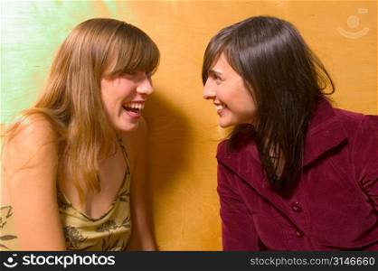 Fashionable Caucasian Girlfriends Sitting Against A Yellow Wall And Laughing At Each Other