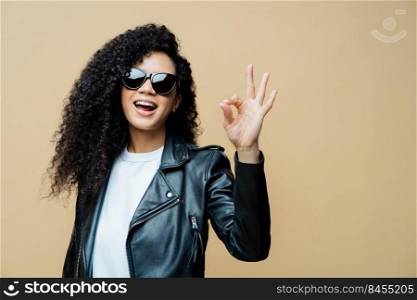 Fashionable curly haired woman tells all is fine, makes okay gesture, says yes to new opportunities, wears trendy sunglasses and black leather jacket, isolated on brown background. Body language