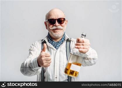 Fashionable elderly man holds the bottle of good alcohol, grey background. Mature senior looking at camera in studio, dude. Elderly man holds the bottle of good alcohol