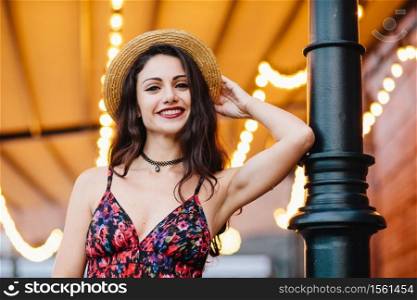 Fashionable elegant woman wearing dress with flower print and straw hat, posing at camera with smile, feeling relaxation, having good mood. People, beauty, lifestyle, facial expression, youth concept