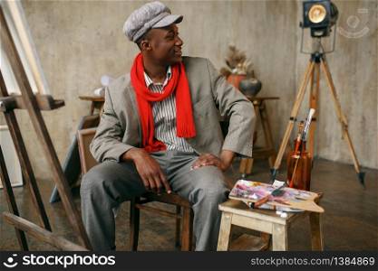 Fashionable male painter in hat and red scarf poses at easel in art studio. Artist draws at his workplace, creative master works in workshop. Painter in hat and red scarf poses at easel