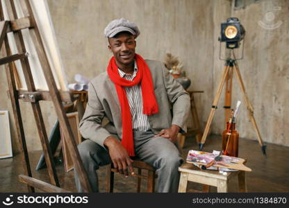 Fashionable male painter in hat and red scarf poses at easel in art studio. Artist draws at his workplace, creative master works in workshop. Painter in hat and red scarf poses at easel