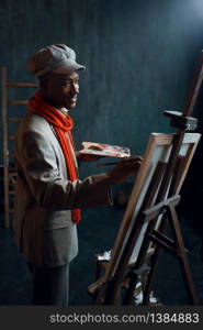 Fashionable male painter with palette poses at easel in art studio. Artist draws at his workplace. Fashionable painter with palette poses at easel