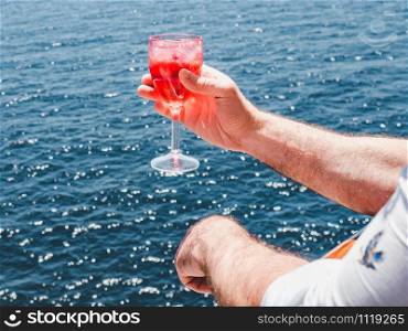 Fashionable man holding a beautiful glass of pink wine on the open deck of a cruise liner against the backdrop of blue sea waves. Side view, close-up. Concept of leisure and travel. Man holding a beautiful glass of pink wine