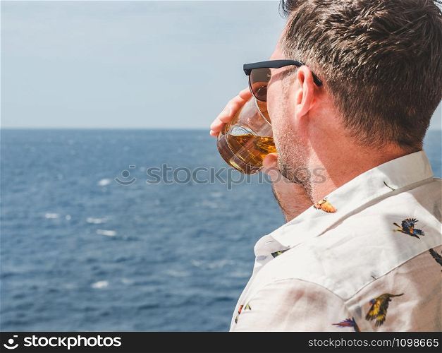 Fashionable man holding a beautiful glass on the open deck of a cruise liner against the backdrop of blue sea waves. Side view, close-up. Concept of leisure and travel. Fashionable man holding a beautiful glass