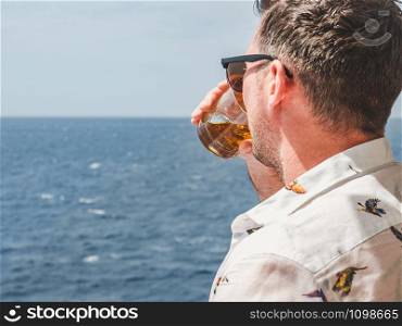 Fashionable man holding a beautiful glass on the open deck of a cruise liner against the backdrop of blue sea waves. Side view, close-up. Concept of leisure and travel. Fashionable man holding a beautiful glass