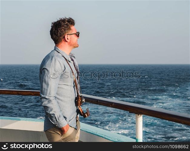 Fashionable man holding a vintage camera on the empty deck of a cruise liner against a background of sea waves. Side view, close-up. Concept of leisure and travel. Man on the empty deck of a cruise liner