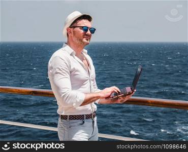 Fashionable man in sunglasses and a sun hat, working on a red laptop on the empty deck of a cruise liner against the background of sea waves. View from the back. Concept of leisure and travel. Fashionable man, working on a red laptop