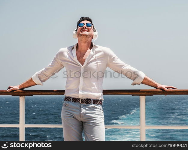 Fashionable man listening to music in white headphones on the empty deck of a cruise liner against the backdrop of the sea waves. Side view, close-up. Concept of technology, recreation and travel. Fashionable man listening to music in white headphones