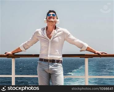Fashionable man listening to music in white headphones on the empty deck of a cruise liner against the backdrop of the sea waves. Side view, close-up. Concept of technology, recreation and travel. Fashionable man listening to music in white headphones