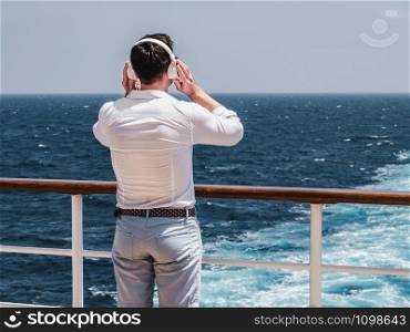 Fashionable man listening to music in white headphones on the empty deck of a cruise liner against the backdrop of the sea waves. Side view, close-up. Concept of technology, recreation and travel. Fashionable man listening to music in white headphones