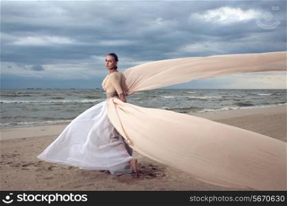 Fashionable model with extremely long dress like a wings. Vogue style. Beautiful model walks at the beach