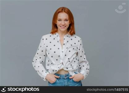 Fashionable positive woman wears polka dot shirt and jeans, keeps hands in pockets, stands self assured against grey background, has happy casual talk with interlocutor. Emotions, fashion concept