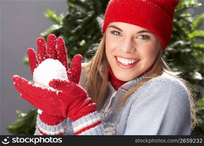 Fashionable Teenage Girl Wearing Cap And Knitwear Holding Snowball In Studio In Front Of Christmas Tree