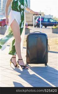 Fashionable woman arriving to new city wearing long dress and high heels, holding her suitcase on wheels admiring town after arrival.. Fashion model traveling to new city