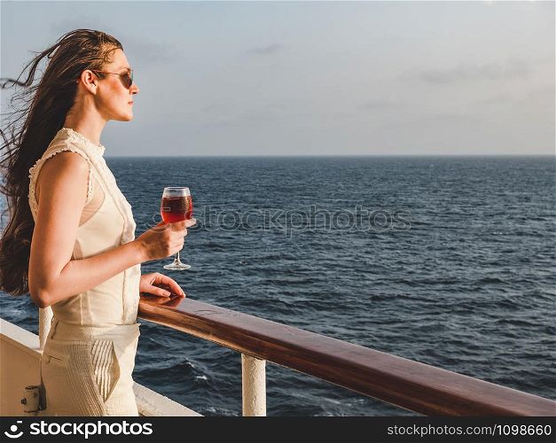 Fashionable woman holding a beautiful glass of pink wine on the open deck of a cruise liner against the backdrop of blue sea waves. Side view, close-up. Concept of leisure and travel. Woman holding a beautiful glass of pink wine