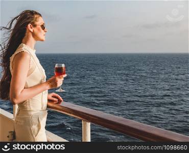 Fashionable woman holding a beautiful glass of pink wine on the open deck of a cruise liner against the backdrop of blue sea waves. Side view, close-up. Concept of leisure and travel. Woman holding a beautiful glass of pink wine