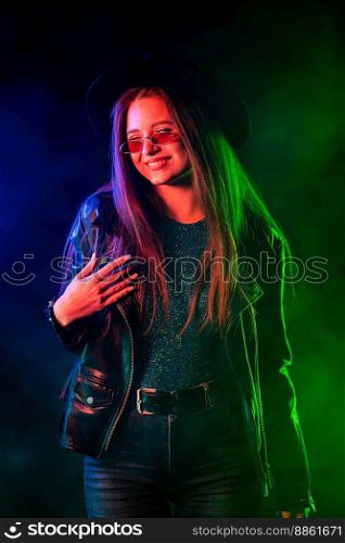 Fashionable woman in neon multi-color light. Smoky background, night party, disco club. Z-generation enigmatic beautiful lady in leather jacket, hat, eyewear. High quality photo. Fashionable woman in neon multi-color light. Smoky background, night party, disco club. Z-generation enigmatic beautiful lady in leather jacket, hat, eyewear