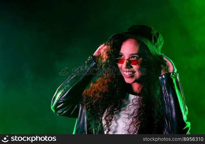 Fashionable woman in neon multi-color light. Smoky background, night party, disco club. Z-generation enigmatic beautiful lady in leather jacket, hat, eyewear. High quality photo. Pretty woman in green neon light. Smoky background, night party, disco club.