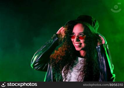 Fashionable woman in neon multi-color light. Smoky background, night party, disco club. Z-generation enigmatic beautiful lady in leather jacket, hat, eyewear. High quality photo. Pretty woman in green neon light. Smoky background, night party, disco club.
