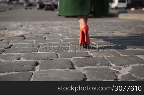 Fashionable woman in orange high heel shoes and stylish emerald green coat walking on cobblestone street to the parked car. Back view of elegant female legs walking on paving stone road closeup. Focus ends on flashing car headlights. Slow motion.