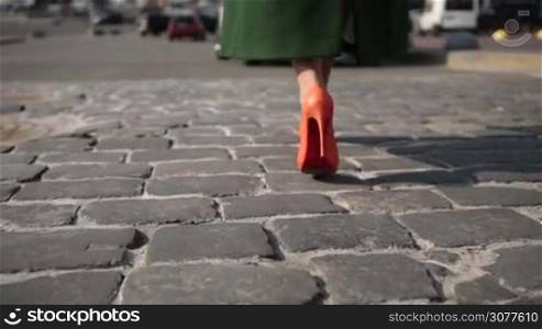 Fashionable woman in orange high heel shoes and stylish emerald green coat walking on cobblestone street to the parked car. Back view of elegant female legs walking on paving stone road closeup. Focus ends on flashing car headlights. Slow motion.