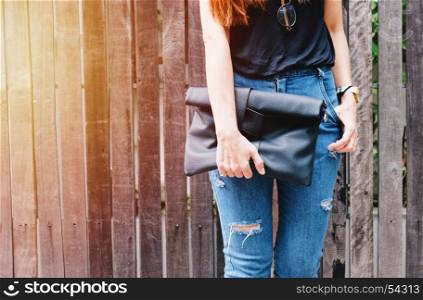 Fashionable woman wearing ripped jeans holding handbag posing at the street with wooden wall, copy space