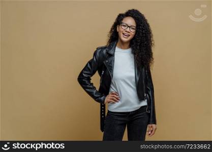 Fashionable woman with glad expression, keeps hand on hip, smiles cheerfully, wears optical glasses, white t shirt, black leather jacket and jeans, ready for walk, poses against beige background