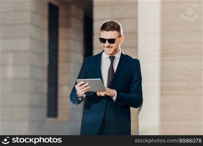 Fashionable young male banker examines future banking agreement, uses modern touch pad, reads important information in internet wears trendy sunglasses and luxury formal suit, smiles gladfully