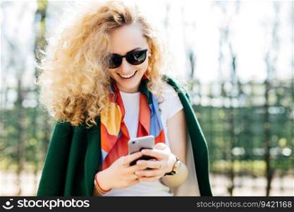 Fashionable young woman in round shades texting friends via social networks, enjoying sunny weather in green park. People, communication, lifestyle.
